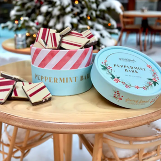 A round tin of peppermint bark chocolate bars sits on a wooden table outdoors, with some pieces broken out and placed in front. The tin is light blue with red-and-white diagonal stripes and gold lettering, and the matching lid rests beside it.