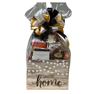 A gift basket with a 'WELCOME HOME' sign, wrapped in decorative paper and tied with a ribbon, containing various food items.